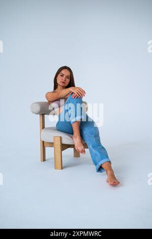 una ragazza in jeans si siede su una sedia con la gamba allungata e le mani addosso Foto Stock