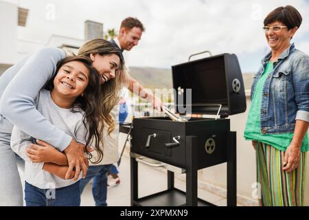Famiglie che fanno barbecue sul tetto di casa - amici multirazziali felici che si divertono a mangiare e cucinare insieme durante il giorno del fine settimana - Estate e cibo Foto Stock