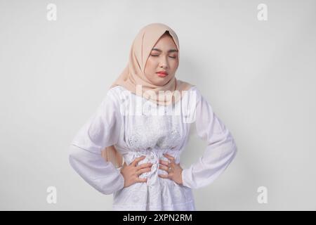 Giovane donna musulmana asiatica in abito bianco e crema velo hijab che tiene il suo stomaco sentendosi affamato durante il digiuno. Concetto Ramadan Foto Stock