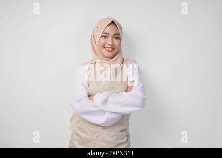 Giovane donna musulmana asiatica che indossa hijab e grembiule color crema sorridente alla macchina fotografica con le braccia incrociate, è piena di fiducia Foto Stock