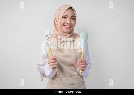 Eccitata giovane donna musulmana asiatica in un velo hijab e grembiule panna sorridente alla macchina fotografica tenendo spatola e utensili da cucina Foto Stock