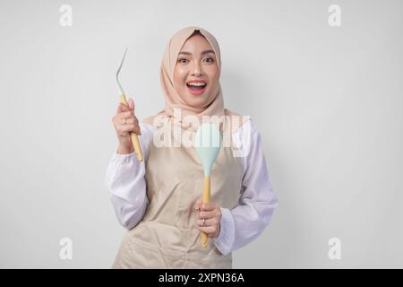 Eccitata giovane donna musulmana asiatica in un velo hijab e grembiule panna sorridente alla macchina fotografica tenendo spatola e utensili da cucina Foto Stock