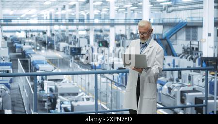 Esperto scienziato industriale che indossa un cappotto bianco, utilizza un computer portatile e si occupa della produzione in una moderna fabbrica di elettronica automatizzata con bracci robotici idraulici Foto Stock