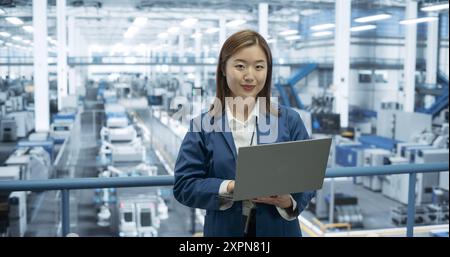 Ritratto di un bellissimo ingegnere della produzione elettronica sorridente, guardando la fotocamera. Giovane donna giapponese che usa il computer portatile per la ricerca online, monitoraggio dei sistemi di intelligenza artificiale Foto Stock