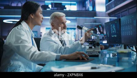 Biotechnology Medical Research Center: Neurologo asiatica femminile che utilizza computer desktop e parla con scienziato caucasico maschio con microscopio. Medici che studiano casi di danno cerebrale, trovano soluzioni. Foto Stock