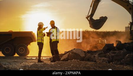 Ripresa cinematografica dell'ora d'oro del cantiere: Ingegnere civile maschio caucasico e urbanista ispanico femminile che parla, usando un tablet. Autocarri, escavatori e caricatori in fase di costruzione di un nuovo blocco di appartamenti Foto Stock