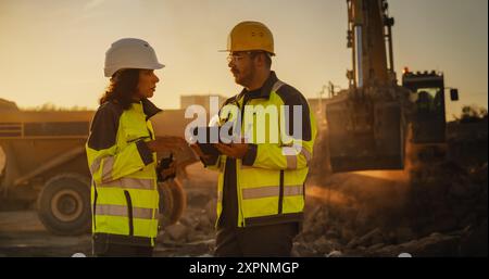 Ripresa cinematografica dell'ora d'oro del cantiere: Ingegnere civile maschio caucasico e urbanista ispanico femminile che parla, usando un tablet. Camion, escavatori, caricatori che lavorano per costruire il New City District. Foto Stock