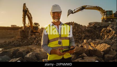 Ingegnere civile con barba di successo che indossa occhiali protettivi e sorride presso la camera on Construction Site in una giornata di sole mentre lavora con macchinari pesanti. Uomo che tiene un tablet, indossa un elmetto, giubbotto di sicurezza. Foto Stock