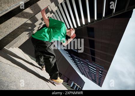 Il Breakdancer in camicia verde si posa sui gradini urbani, sfidando la gravità con una mossa elegante contro l'architettura moderna della città. Foto Stock
