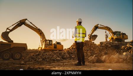 Urban Planner maschio caucasico indossa occhiali protettivi e utilizza tablet in cantiere in Una giornata di sole. Uomo che ispeziona Building Progress. Carico di materiali su dumper industriali da parte dell'escavatore Foto Stock