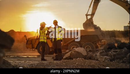 Ripresa cinematografica dell'ora d'oro del cantiere: Ingegnere civile maschio caucasico e urbanista ispanico femminile che parla, usando un tablet. Autocarri, escavatori, caricatori che lavorano per la costruzione di blocchi di appartamenti Foto Stock