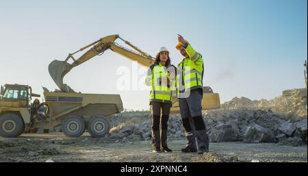 Ispettore femminile ispanico che parla con il responsabile dello sviluppo del territorio maschile caucasico con tablet on Construction Site of Real Estate Project. Escavatori industriali in preparazione per la posa delle fondamenta. Foto Stock