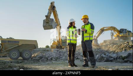 Ispettore femminile ispanico che parla con il responsabile dello sviluppo del territorio maschile caucasico con tablet on Construction Site of Real Estate Project. Escavatori industriali in preparazione per la posa di fondamenta per l'edilizia. Foto Stock