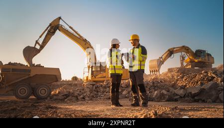 Ispettore femminile ispanico che parla con il responsabile dello sviluppo del territorio maschile caucasico con tablet on Construction Site of Real Estate Project. Escavatori in preparazione per la posa delle fondamenta. Caldo giorno di sole Foto Stock