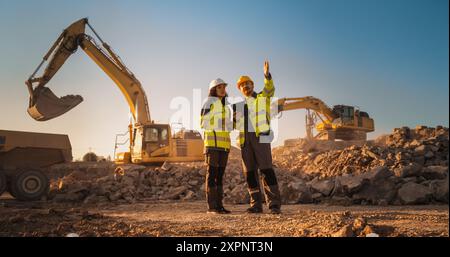 Ispettore femminile ispanico che parla con il responsabile dello sviluppo del territorio maschile caucasico con tablet on Construction Site of Real Estate Project. Escavatori in preparazione per la posa di fondamenta edili. Caldo giorno di sole Foto Stock