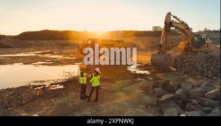 Drone aereo Shot of Construction Site on Sunny Evening: Gli escavatori industriali scavano rocce e le caricano nel camion. Ingegnere civile e architetto osservando il processo, discutendo del progetto immobiliare Foto Stock