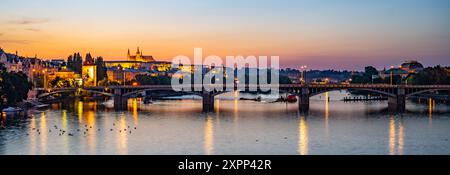 Il ponte Jirasek attraversa il fiume Moldava a Praga, con il castello di Praga illuminato sulla riva opposta. Il sole che tramonta proietta una calda luce sul paesaggio cittadino, creando una scena serena e pittoresca. Foto Stock