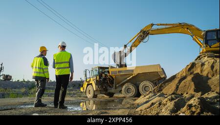 Un investitore immobiliare e Project Manager uomo caucasico che parla sul sito di costruzione di un nuovo blocco di appartamenti. Colleghi che discutono di progressi, affari. Sabbia di carico dell'escavatore in un dumper industriale. Foto Stock