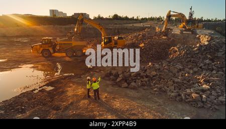 Drone aereo Shot of Construction Site on Sunny Evening: Escavatori industriali che scavano rocce e le caricano su un camion. Ingegnere e architetto che osservano il processo, discutono del progetto immobiliare Foto Stock