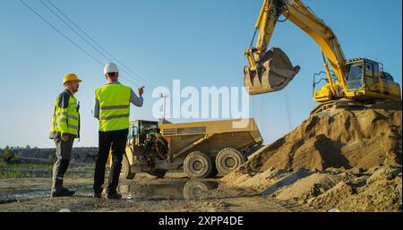 Un investitore immobiliare e Project Manager uomo caucasico parla sul sito di costruzione di Apartment Block. Colleghi che discutono del progresso degli edifici, affari. Sabbia di carico dell'escavatore in un dumper industriale. Foto Stock