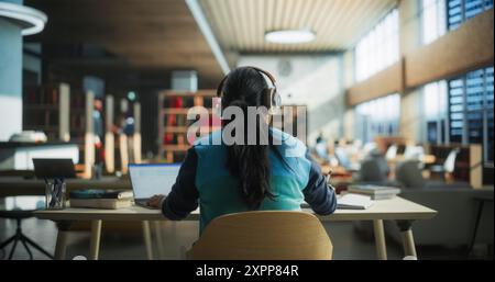 Studentessa che indossa le cuffie mentre lavora alle esercitazioni universitarie in una biblioteca pubblica. Donna seduta dietro una scrivania, che utilizza un computer portatile e scrive appunti nel notebook. Riprese dal retro Foto Stock