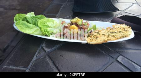 Antipasto alla tartare di tonno con foglie di lattuga e pane piatto al parmigiano su un piatto bianco oblungo, seduto su un bancone di pietra nera. Foto Stock