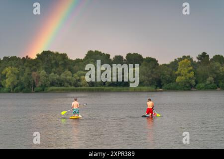 Due tizi su tavole SUP, essendo al centro del fiume, nuotano fino alla riva opposta, sopra la quale è visibile un arcobaleno. Foto Stock