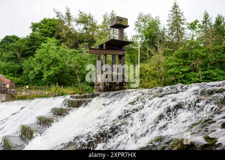 L'acqua scorre su una cascata con una struttura rustica in legno e metallo e lussureggianti alberi verdi sullo sfondo. Foto Stock