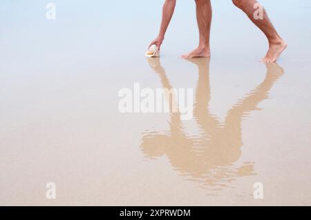 Uomo sulla sabbia bagnata di prelevare un guscio e la sua riflessione su di essa. Foto Stock