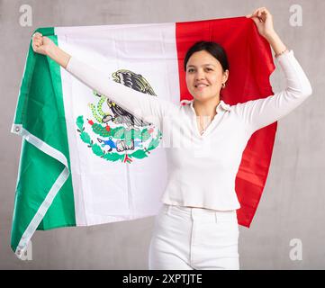 La giovane donna positiva e affidabile detiene la grande bandiera nazionale del Messico con un sorriso fiducioso che mostra i denti Foto Stock