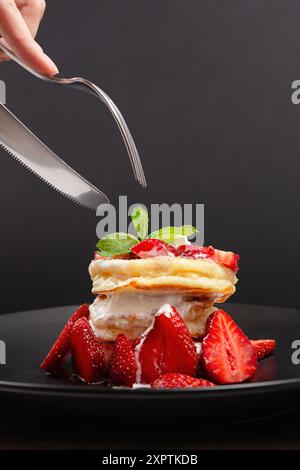 Primo piano di mano con coltello e forchetta per tagliare una pila di soffici pancake ricoperti di fragole fresche e panna montata. Foto Stock