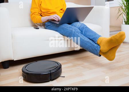 Persona in maglione giallo che utilizza un computer portatile su un divano bianco, gambe allungate, con un aspirapolvere robot sul pavimento di legno. Foto Stock