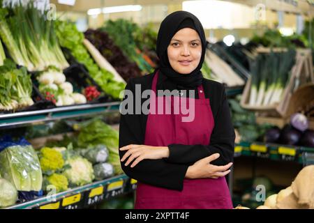 Ritratto di amichevole venditore femminile in hijab e grembiule nella sezione della drogheria del supermercato Foto Stock