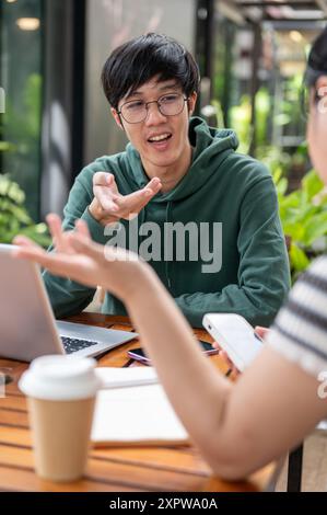 Un giovane asiatico in una felpa verde con cappuccio sta parlando, discutendo di lavoro con il suo amico seduto a un tavolo all'aperto. brainstorming, consulenza, lavoro di squadra Foto Stock