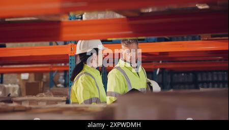 Logistica, colleghi e camminate in fabbrica, ispettore e inventario delle scorte per la spedizione e le conversazioni. Comunicazione, casco e persone in magazzino Foto Stock