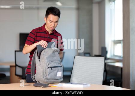 Un giovane studente universitario sta imballando vari oggetti nel suo zaino in una classe, tra cui il suo computer e i libri Foto Stock