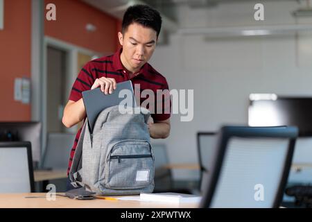 Un giovane studente universitario sta imballando vari oggetti nel suo zaino in una classe, tra cui il suo computer e i libri Foto Stock