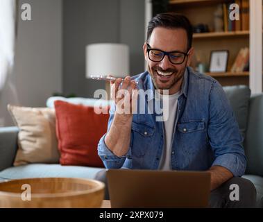 Uomo d'affari sorridente che controlla le e-mail sul computer portatile e parla sullo smartphone in ufficio. Libero professionista che utilizza un computer wireless e invia voic Foto Stock