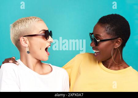 Donne, wow e occhiali da sole con sorpresa, insieme e incollati su sfondo blu come amici. Le donne, lo shock e il sorriso sono allegri nella diversità Foto Stock