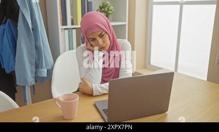 Una donna premurosa che indossa un hijab siede in un ufficio domestico con un notebook, riflettendo un momento di contemplazione. Foto Stock