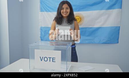 Giovane donna che vota in casa con sfondo bandiera argentina, sorridendo nella sala del collegio elettorale tenendo una busta vicino a un ballottaggio. Foto Stock