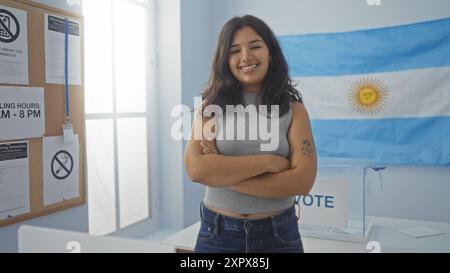 Giovane donna con le braccia incrociate che sorride in una sala votante al coperto con una bandiera argentina e istruzioni di voto sul muro che indicano un set elettorale Foto Stock