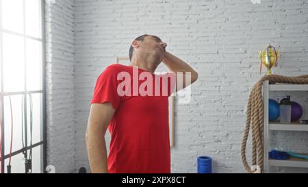 Un uomo ispanico di mezza età con una camicia rossa viene visto massaggiarsi il collo in una palestra interna illuminata, circondata da attrezzature per l'allenamento contro un bri bianco Foto Stock