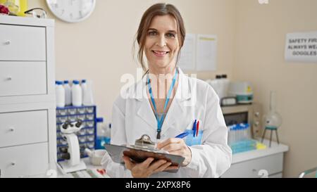 Una donna sorridente di mezza età in un camice da laboratorio prende appunti in un laboratorio, rappresentando la professionalità nel settore sanitario. Foto Stock