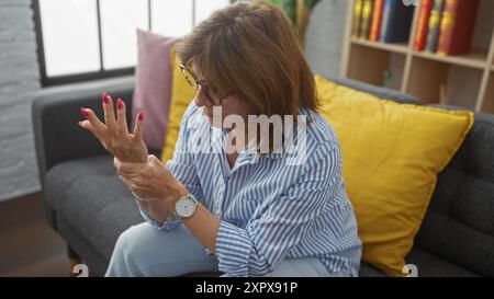 Una donna di mezza età esamina il disagio alla mano in un salotto accogliente, raffigurando dolore o artrite Foto Stock