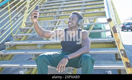 Uomo afroamericano che scatta selfie con lo smartphone sulle scale della città all'aperto indossando le cuffie. Foto Stock