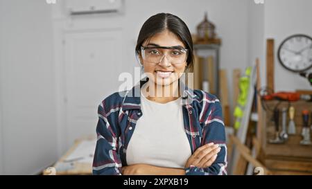 Una giovane donna indiana con capelli lunghi e occhiali di sicurezza sorride con fiducia in un laboratorio di falegnameria ben attrezzato. Foto Stock