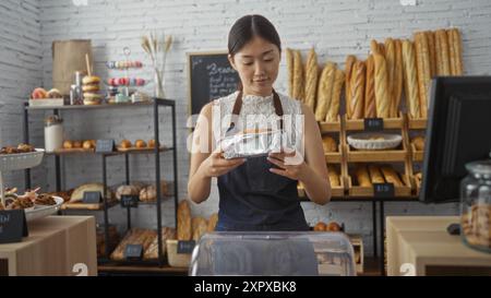 Giovane donna cinese che tiene il pane in una panetteria con scaffali pieni di prodotti appena sfornati sullo sfondo Foto Stock