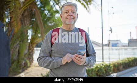 Uomo dai capelli d'argento con zaino e smartphone in piedi su una strada con palme e ambientazione urbana sullo sfondo Foto Stock