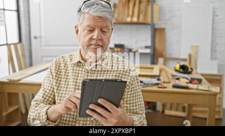 Un uomo anziano con capelli grigi concentrato su un tablet in un'officina di legno circondata da utensili e banco da lavoro. Foto Stock